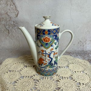 May include: A white ceramic teapot with a long spout and handle, featuring a colorful floral and landscape design. The design includes orange, blue, and green accents. The lid is also decorated with floral patterns. The teapot is placed on a cream-colored lace doily.