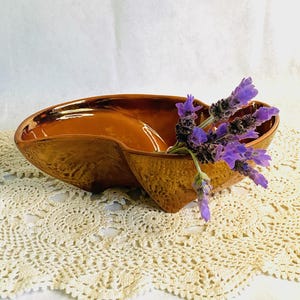 May include: A two-sectioned, brown ceramic bowl with a textured exterior and glossy interior. The bowl is filled with purple lavender sprigs and sits on a cream-colored, crocheted doily. The bowl's shape is reminiscent of a heart or a stylized leaf.