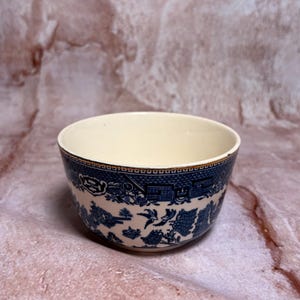 May include: A ceramic bowl with a cream-colored interior and a blue and white patterned exterior. The pattern features a landscape design with buildings, trees, and birds. The bowl has a gold rim and is set against a pink and brown background.