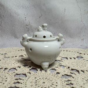 May include: A white ceramic incense burner with a rounded body, three small feet, and a lid with a decorative finial. The burner has several small holes for releasing incense smoke and two decorative handles.