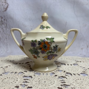 May include: Cream-colored ceramic sugar bowl with a lid and two handles. The bowl is decorated with a floral pattern in orange, purple, green, and blue. The sugar bowl is sitting on a cream-colored lace doily.