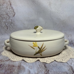May include: Cream-colored ceramic casserole dish with lid and handles. The dish features a yellow floral design and a green and cream-colored knob on the lid. It sits on a cream-colored lace doily.