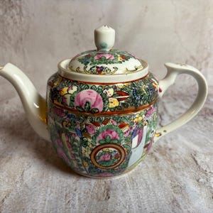 May include: A white porcelain teapot with a lid, featuring a colorful floral and figural design. The teapot has a curved spout and handle, and the lid is topped with a small knob. The design includes pink, green, and yellow flowers.