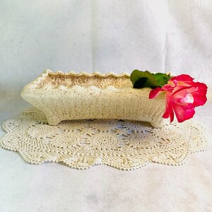 May include: A cream-colored rectangular planter with a textured surface and scalloped rim. A red and pink rose with a green leaf rests on the planter. The planter sits on a cream-colored, crocheted doily.