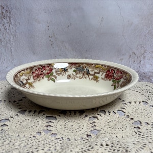 May include: Cream-colored ceramic bowl with a floral pattern around the rim. The rim has a scalloped edge. The floral design features red, brown, and green elements. The bowl is set on a cream-colored lace doily.