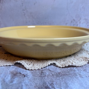 May include: A light yellow pie dish with a scalloped edge and a ridged exterior. The dish is sitting on a cream-colored, crocheted doily, against a mottled gray and blue background. A classic kitchenware item.
