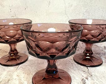 May include: Three vintage amethyst-colored glass dessert bowls. Each bowl has a diamond pattern and sits on a stem with a circular base. The bowls are arranged on a light-colored surface.