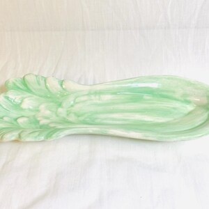 May include: A ceramic serving dish in the shape of a stylized lettuce leaf. The dish is painted in shades of light green and white, with a glossy finish. The leaf-shaped dish is ideal for serving appetizers or small dishes.