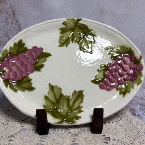 May include: Oval white ceramic serving platter with embossed purple grapes and green leaves. The platter is supported by two dark brown wooden stands. A decorative piece for serving food.