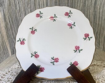 May include: White porcelain plate with a scalloped edge and gold trim. The plate is decorated with pink rose floral patterns. The plate is displayed on a dark brown wooden stand.