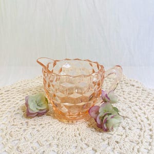 May include: A vintage, peach-colored glass creamer with a diamond pattern. The creamer has a handle and a spout. It is sitting on a cream-colored, crocheted doily, with a few small flowers.