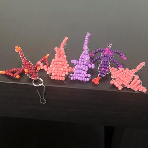 Pu&ograve; includere: Una collezione di ciondoli fatti a mano con perline di vari colori, tra cui rosso, rosa e viola. I ciondoli hanno la forma di figure astratte e sono attaccati a portachiavi color argento. I ciondoli sono esposti su una superficie scura.