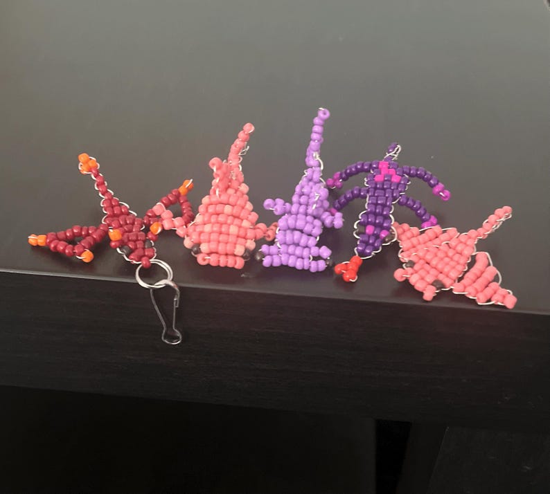 Pu&ograve; includere: Una collezione di ciondoli fatti a mano con perline di vari colori, tra cui rosso, rosa e viola. I ciondoli hanno la forma di figure astratte e sono attaccati a portachiavi color argento. I ciondoli sono esposti su una superficie scura.