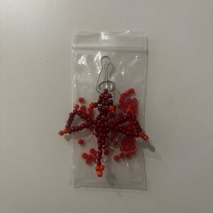 Pu&ograve; includere: Un ornamento a forma di libellula di perline in un sacchetto di plastica trasparente. La libellula &egrave; fatta di piccole perline rosso scuro con accenti arancioni sulle ali e sulla testa. Una clip in metallo color argento &egrave; attaccata alla parte superiore dell'ornamento.