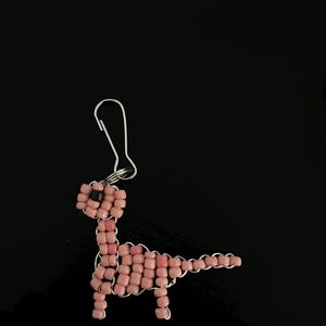 Pu&ograve; includere: Orecchino a forma di dinosauro fatto a mano con perline rosa e gancio color argento. Il dinosauro &egrave; realizzato con piccole perline quadrate e filo metallico, con una perlina nera per l'occhio. Su sfondo nero.