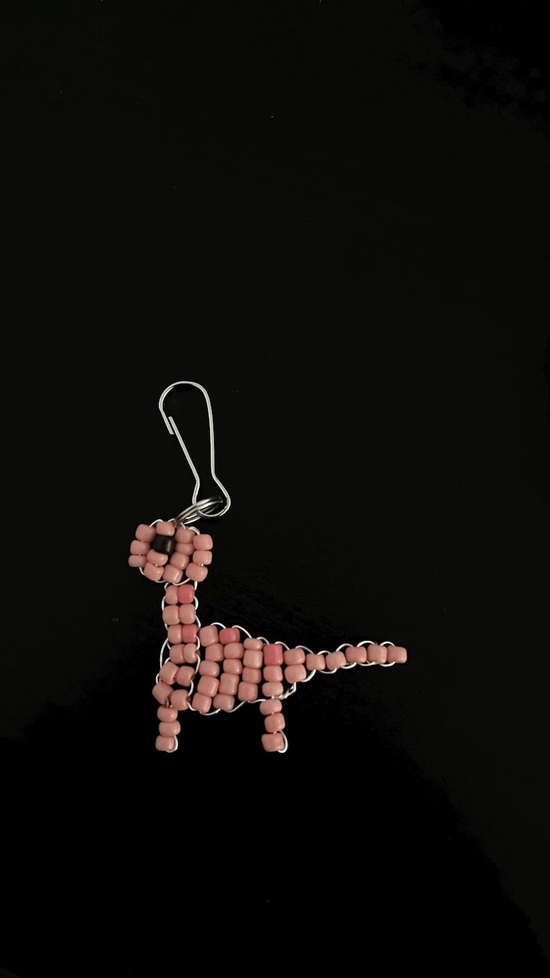 Pu&ograve; includere: Orecchino a forma di dinosauro fatto a mano con perline rosa e gancio color argento. Il dinosauro &egrave; realizzato con piccole perline quadrate e filo metallico, con una perlina nera per l'occhio. Su sfondo nero.