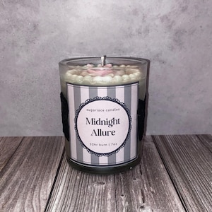 May include: A glass candle jar with a label that reads "Midnight Allure." The candle has a white and pink wax top with a silver wick. The label has a striped design with black lace accents. The candle is 7oz and has a 30hr burn time.