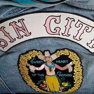 May include: A denim jacket with two embroidered patches. The top patch reads "SIN CITY" in pink lettering. The bottom patch features a woman in a cowboy hat with the text "SWEET HEART OF THE RODEO".