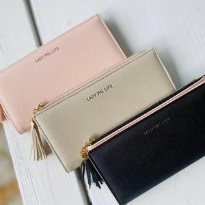 Può includere: Tre portafogli rettangolari in rosa chiaro, beige e nero, ciascuno dotato di una cerniera dorata con un tirante a nappa abbinato e il testo LADY PAL LIFE impresso in lettere dorate sulla parte anteriore di ogni singolo portafoglio.