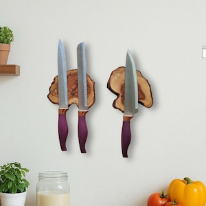 Puede incluir: Tres cuchillos de cocina con mangos morados y hojas plateadas se exhiben en un soporte de madera. El soporte es de madera clara de forma orgánica. Los cuchillos están montados en una pared blanca.