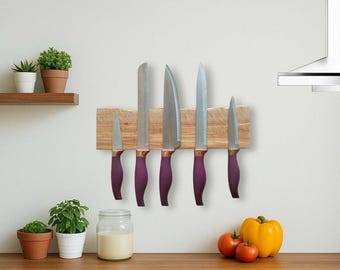 Può includere: Un portacoltelli in legno con sei coltelli in acciaio inossidabile. I coltelli hanno manici viola e sono montati su un blocco di legno chiaro. La scena della cucina comprende piante e verdure.