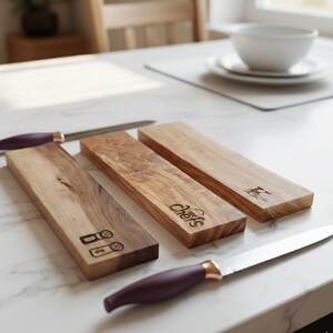 Könnte beinhalten: Messerblöcke aus Holz mit eingravierten Designs und zwei Messern mit lila Griffen und kupferfarbenen Akzenten. Die Blöcke tragen das Wort "Chefs" und andere eingravierte Symbole. Die Messer und Blöcke liegen auf einer weißen Marmoroberfläche.