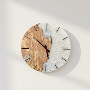 Puede incluir: Un reloj de pared redondo con un diseño único. La mitad de la esfera del reloj es de madera clara con un patrón de vetas natural, mientras que la otra mitad es blanca con una apariencia texturizada. Las manecillas y los marcadores negros completan el diseño.