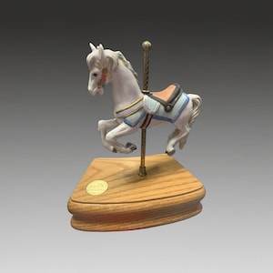 Può includere: Statuetta di cavallo da giostra bianca con sella azzurra e beige, montata su base in legno. Il cavallo ha criniera e coda fluenti ed è attaccato a un palo dorato. Un piccolo emblema dorato è sulla base.