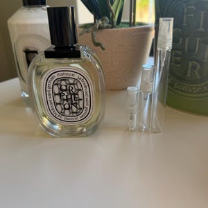 May include: A clear glass perfume bottle with a black cap and a white label with black text. The bottle is labelled "Orpheon" and "diptyque". Several small, clear glass vials are next to the bottle. A white bottle with a black pump is in the background.