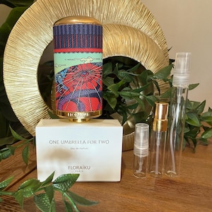 May include: A perfume bottle with a vibrant umbrella design, a gold cap, and the text "ONE UMBRELLA FOR TWO". Several clear spray bottles and a box with the brand name "FLORAIKU PARIS" are also visible.