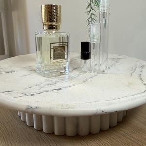 May include: A perfume bottle with a gold cap and the text "EX NIHILO" and "SPICY MUSK" sits on a white marble cake stand. Several clear sample spray bottles are next to the perfume bottle. The cake stand is on a wooden surface.