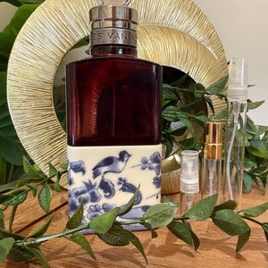 May include: A dark red perfume bottle with a silver cap and the text "ES VAN M". The bottle sits on a white base with a blue and white bird design. Several small spray bottles are also visible.