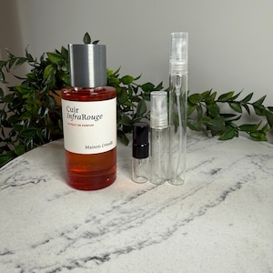 May include: A perfume bottle with a silver cap and a label that reads "Cuir InfraRouge Extrait de Parfum Maison Crivelli." Several clear spray bottles of varying sizes are next to the perfume bottle. The perfume is a reddish-orange colour.