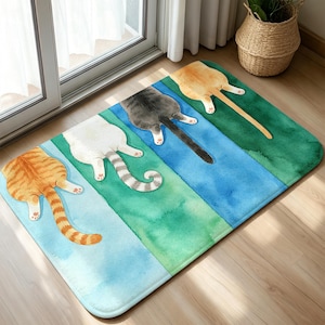 May include: A rectangular floor mat featuring a watercolor illustration of four cat hindquarters in a row. The cats are orange tabby, white with gray stripes, solid gray, and solid orange, each positioned on a distinct blue or green panel.