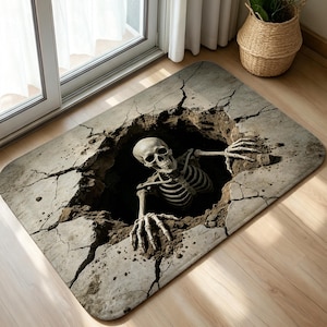 May include: A rectangular floor mat featuring a 3D-style graphic of a human skeleton emerging from a dark, cracked hole in a concrete surface. The design displays realistic textures of broken stone, dirt, and bone in shades of gray and brown.