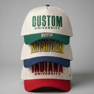 Peut inclure: Pile de quatre casquettes de baseball avec des visières de couleurs différentes et du texte. La casquette supérieure est crème avec une visière verte et le mot "CUSTOM" en vert. La deuxième casquette a une visière verte et le mot "MICHIGAN" en jaune. La troisième casquette a une visière bleue et le mot "INDIANA" en rouge.