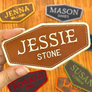 May include: A collection of personalized name patches in various colors and shapes. The patches feature names like "Jessie Stone," "Jenna Williams," and "Mason Dawes" embroidered in white thread. The patches are brown, black, red, green, and orange.