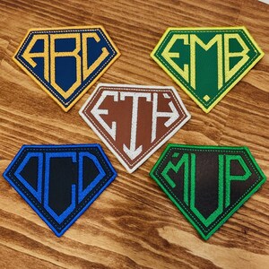 May include: Five embroidered patches in a diamond shape, each featuring a different set of initials. The patches have a superhero-like design with various color combinations, including blue and gold, green and yellow, brown and white, blue and black, and green and black.