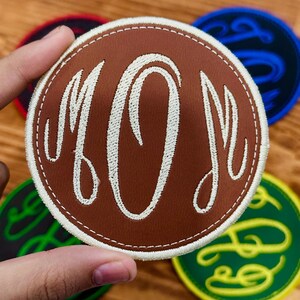 May include: A brown, circular patch with the embroidered word "MOM" in white script. The patch has a white stitched border. Other patches with script lettering in blue, green, and red are visible in the background.