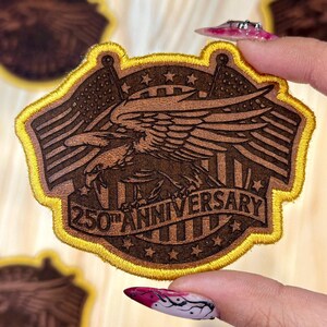May include: A brown patch with a gold border featuring an eagle, American flags, and the text "250th Anniversary." The patch is held by a hand with a decorated fingernail. Other patches are visible in the background.