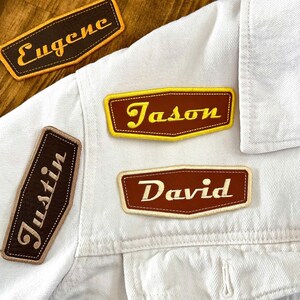 May include: Close-up of a white denim jacket with several embroidered name patches. The patches are brown with yellow or cream-colored text, featuring names like "Eugene", "Jason", "David", and "Justin". The patches have a retro, vintage aesthetic.