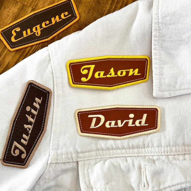 May include: Close-up of a white denim jacket with several embroidered name patches. The patches are brown with yellow or cream-colored text, featuring names like "Eugene", "Jason", "David", and "Justin". The patches have a retro, vintage aesthetic.