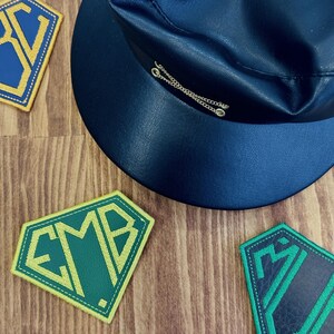 May include: A navy blue hat with a silver chain detail and several embroidered patches. The patches are diamond-shaped with green and yellow lettering. The hat is made of a smooth, dark material.