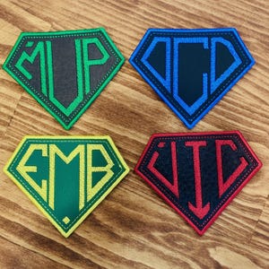 May include: Four diamond-shaped embroidered patches with initials. The patches are green, blue, yellow, and red, each with a different set of initials in a contrasting color. The patches are on a wooden surface.