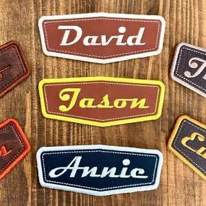 May include: Embroidered name patches in brown, yellow, and navy blue. The patches feature names like "David", "Jason", and "Annie" in white or yellow script. Each patch has a contrasting border and is set against a wooden background.
