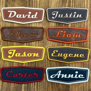 May include: A collection of personalized name patches in various colors. Each patch features a name embroidered in a contrasting color, such as "David" in white on a brown patch, and "Annie" in white on a navy patch. The patches have a stitched border.