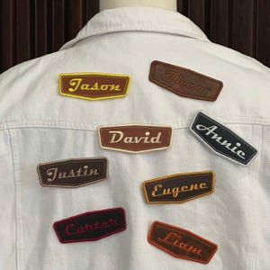 May include: A white denim jacket with several embroidered name patches. The patches feature names like "Jason," "David," and "Annie" in various colors and fonts. The patches are in a retro style.