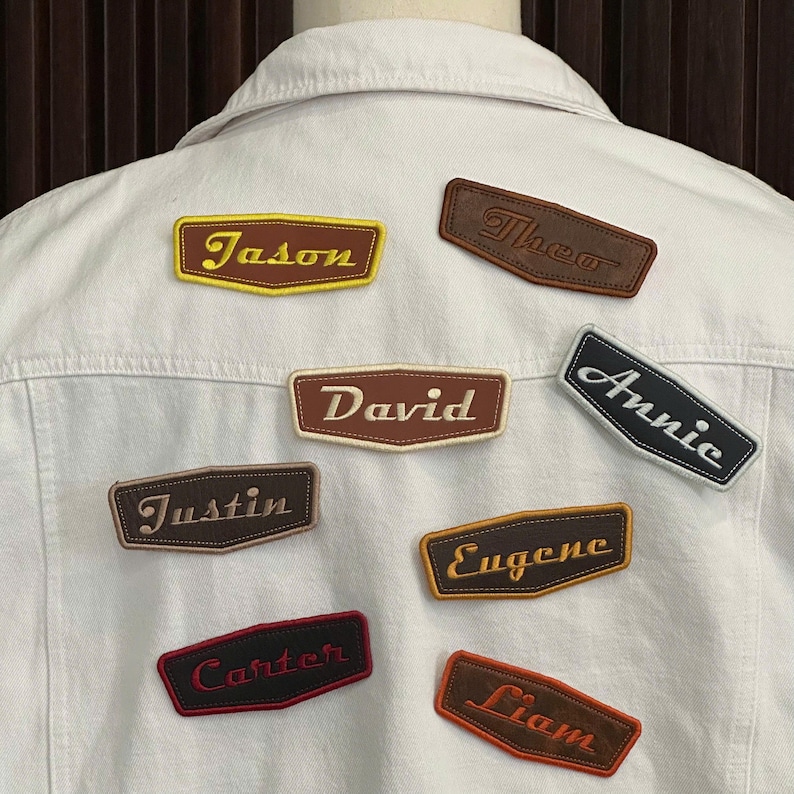May include: A white denim jacket with several embroidered name patches. The patches feature names like "Jason," "David," and "Annie" in various colors and fonts. The patches are in a retro style.