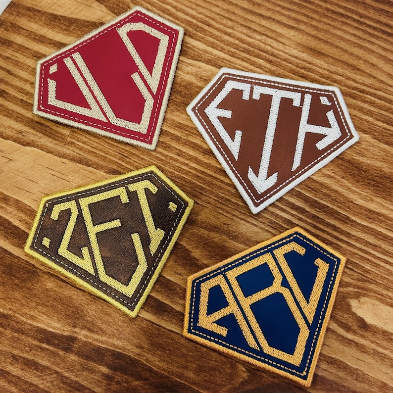 May include: Four diamond-shaped embroidered patches with different color schemes. The patches feature initials in a bold, stylized font. Colors include red, brown, yellow, and blue, with white or yellow stitching. The patches are on a wooden surface.