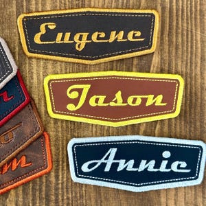 May include: A collection of personalized name patches. The patches are rectangular with a curved bottom edge. The names "Eugene", "Jason", and "Annie" are embroidered in a contrasting color on each patch. The patches have a stitched border.